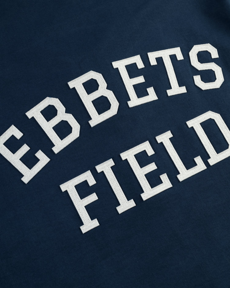 Close-up of white varsity-style EBBETS FIELD lettering on the Ebbets Field Cotton Grounds Crew Jacket by Ebbets Field Flannels—a dark blue cotton piece ideal for spring or as a workwear staple.