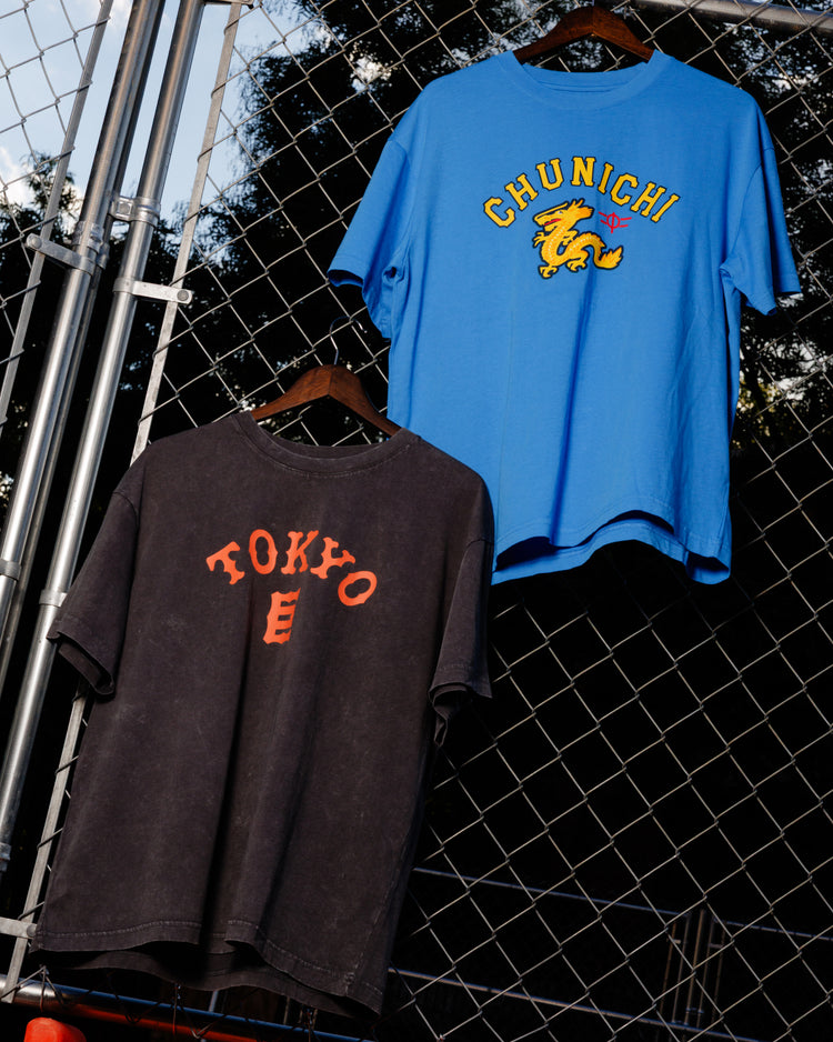 Two graphic tees hang on a fence: a blue CHUNICHI shirt with a yellow dragon, and an Ebbets Field Tokyo Giants Pigment-Dyed Short Sleeve Graphic T-Shirt in black with red letters. Trees and sky are in the background.