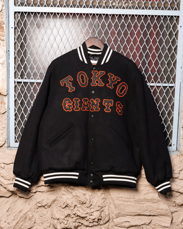The Ebbets Field Tokyo Giants All-Wool Varsity Jacket, featuring bold orange lettering, hangs on a hanger against a wire mesh and stone wall backdrop, honoring Japanese baseball heritage.