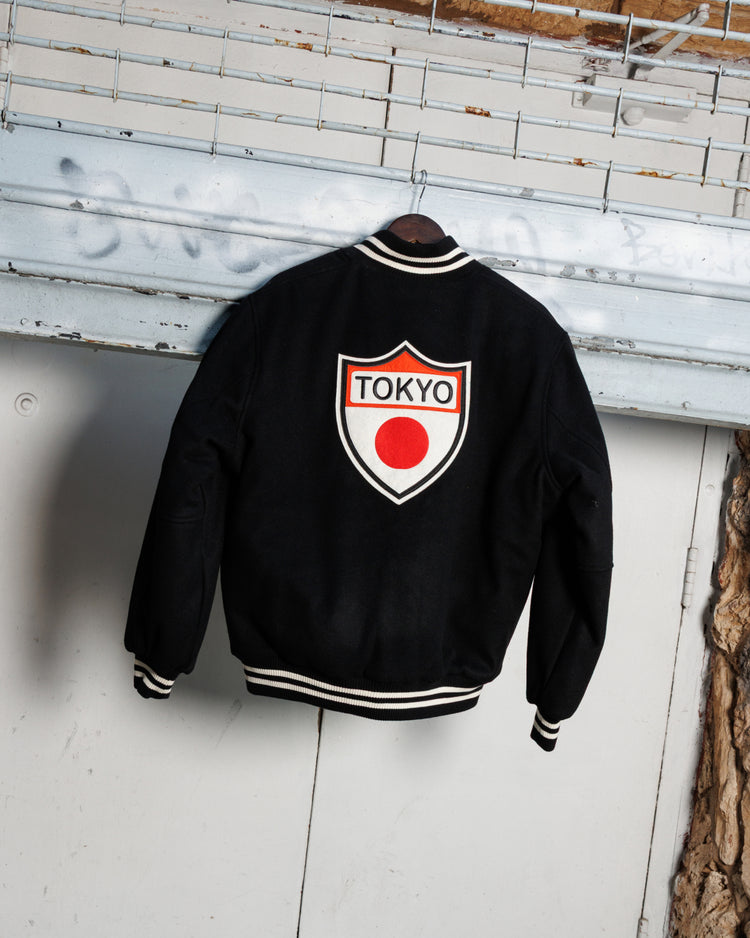 The Ebbets Field Tokyo Giants All-Wool Varsity Jacket in black with white trim hangs on a hanger, showcasing a shield patch, “TOKYO,” and red circle on the back against an industrial metal door backdrop.