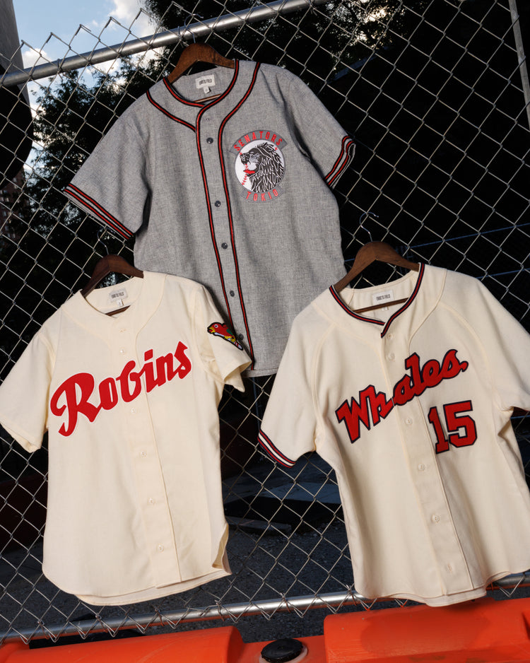 Three vintage-style baseball jerseys hang on a fence, including the Ebbets Field Shochiku Robins 1950 Authentic Wool Home Jersey, each featuring red and black accents on cream or gray fabric.