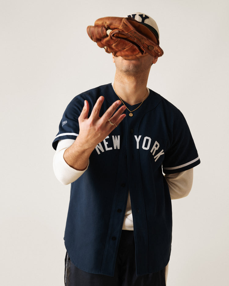 Wearing an Ebbets Field New York Black Yankees Cotton Twill Baseball Jersey and cap, a person tosses a brown baseball glove in the air, partially obscuring their face against a plain light background.