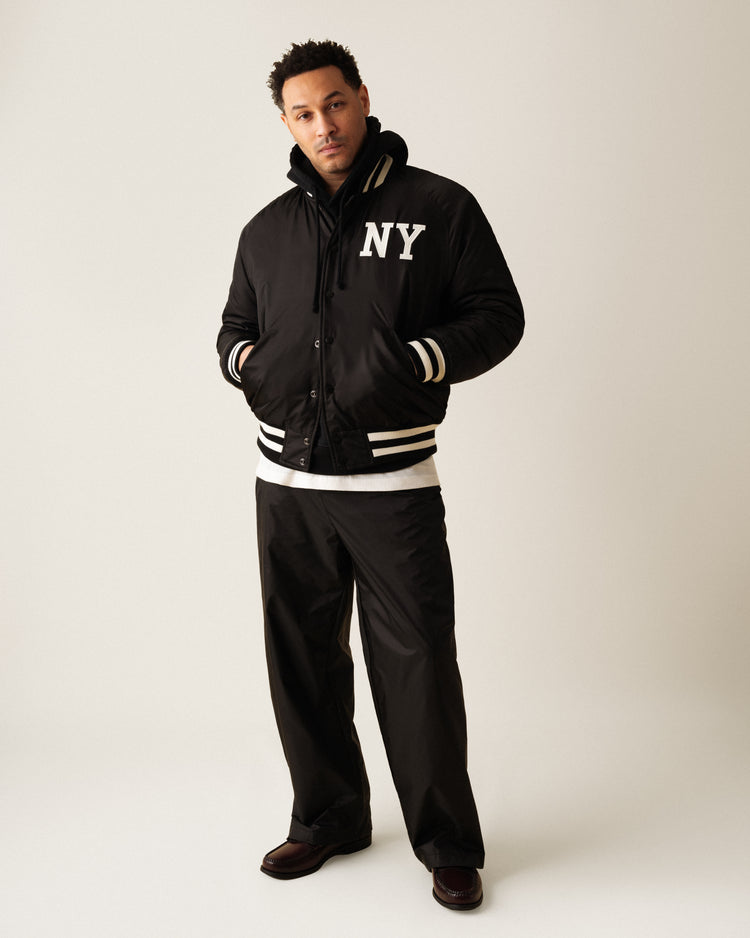 A man stands in a studio wearing the Ebbets Field New York Black Yankees Satin Varsity Jacket, paired with black wide-leg pants, a white shirt, and dark shoes. His hands are in his pockets as he looks confidently at the camera.