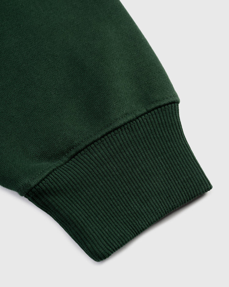 Close-up of the Ebbets Chest Logo Heavyweight Cotton Terry Pullover Hoodie sleeve by Ebbets Field Flannels, featuring a dark green vintage look and ribbed cuff on a light gray background.