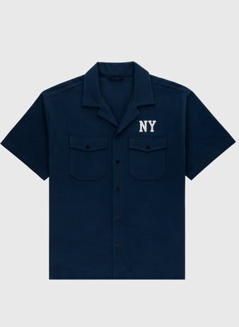 The Ebbets Field Flannels Cotton Linen Grounds Crew Short Sleeve Workshirt in navy blue features two button-flap chest pockets and NY in white above the left pocket. Shirt is displayed flat on a light gray background.