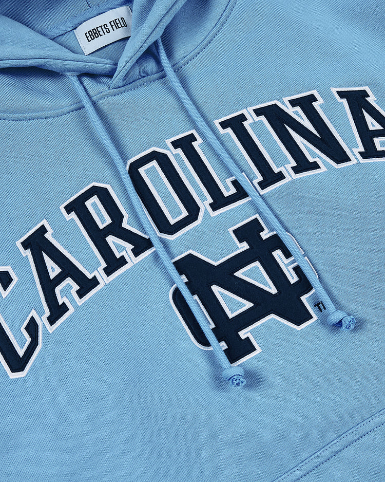 Close-up of the University of North Carolina Heavyweight Cotton Terry Pullover Hoodie by Ebbets Field in light blue, featuring CAROLINA and a large interlocking NC logo on the front, with an Ebbets Field inner neck label.
