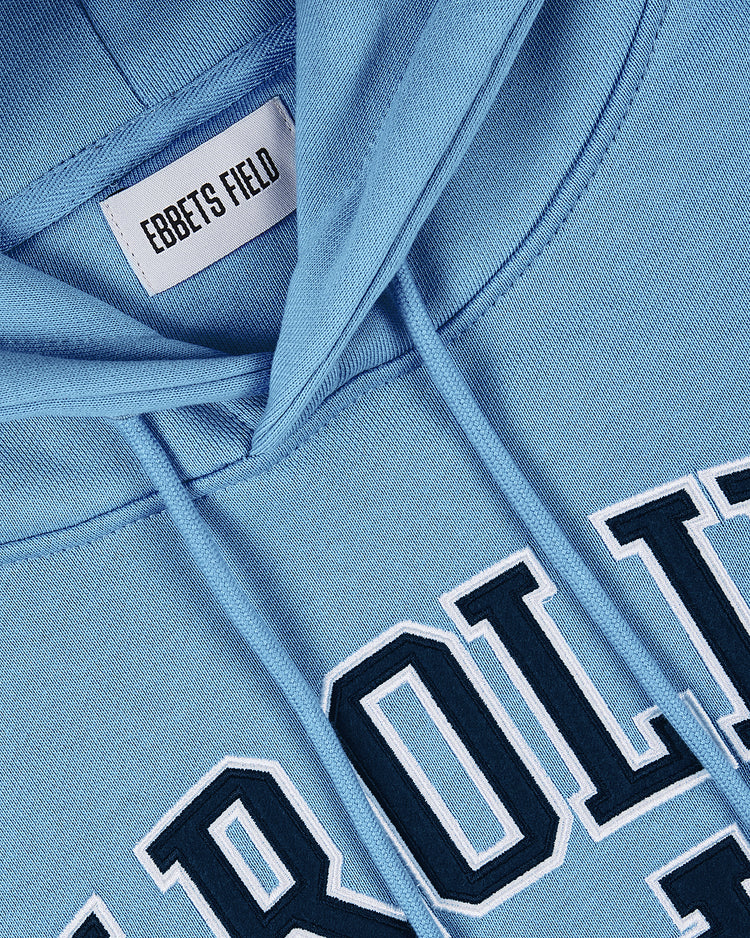 The University of North Carolina Heavyweight Cotton Terry Pullover Hoodie by Ebbets Field features a light blue French terry build, visible brand tag on the collar, and partial embroidered CAROLINA in white and navy for a vintage UNC look.