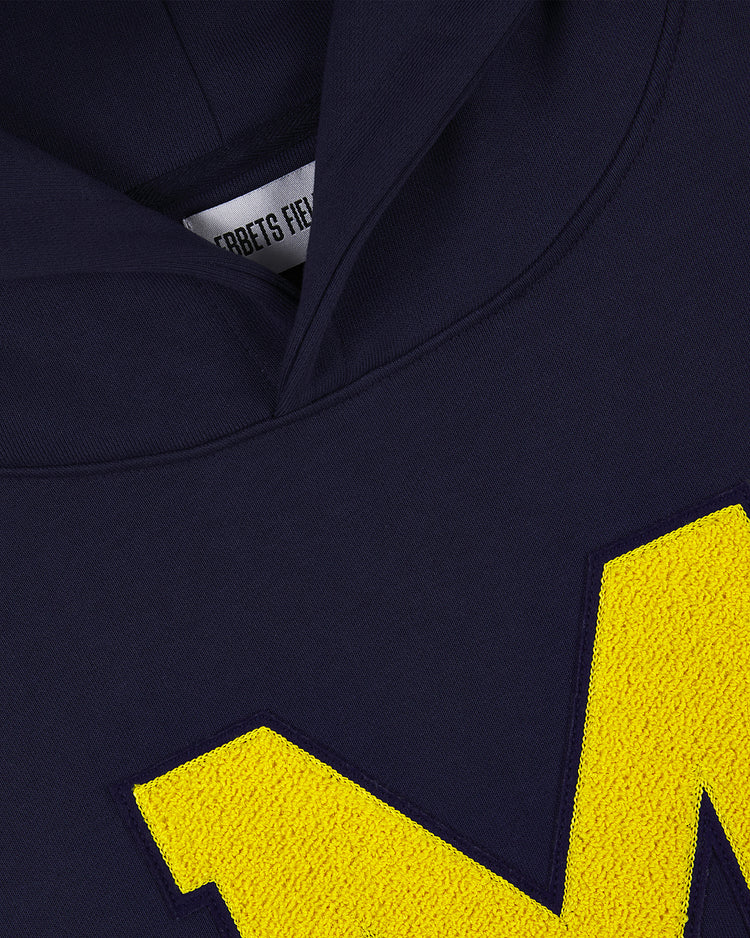 Close-up of the MICHIGAN HOODIE NAVY by Ebbets Field, showcasing a large textured yellow M on the front and a partially visible inside tag with the brand name.
