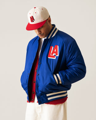 A person wearing a blue varsity jacket with an LA patch, a red and white cap featuring LA, a red shirt, and white pants, looking down with hands in pockets against a plain background.