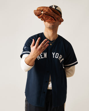A person wearing a New York baseball jersey and cap tosses a brown baseball glove in the air, partially obscuring their face against a plain background.