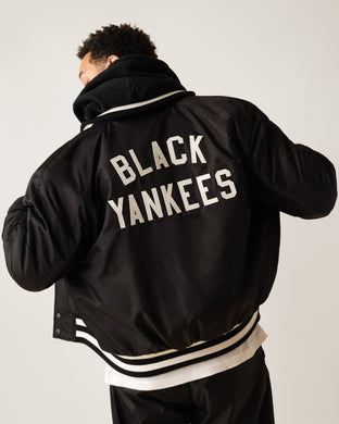 A person wearing a black and white varsity jacket with BLACK YANKEES written in large white letters on the back, standing against a plain light background and facing away from the camera.