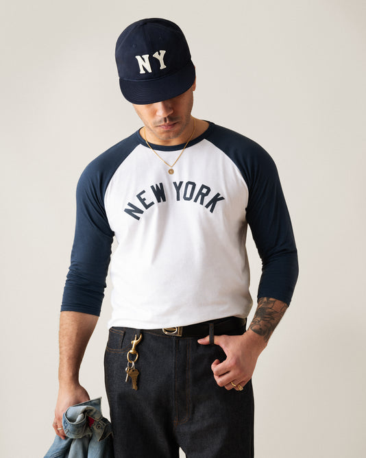 A man wearing a navy and white “New York” baseball shirt and an “NY” cap stands looking down, holding a denim jacket and keys in one hand. He wears dark jeans, a necklace, and has tattoos on his left arm.