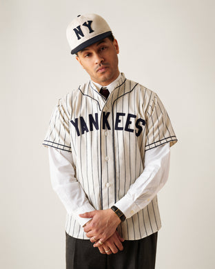 A man wearing a vintage-style New York Yankees baseball jersey and cap, with a white dress shirt and tie underneath, stands with his hands clasped and head tilted slightly to the side against a plain background.