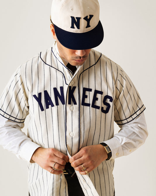 A person wearing a Yankees cap and vintage pinstripe Yankees jersey, buttoning up the shirt while looking down, with a plain background.