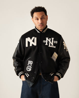 A man stands facing forward, wearing a black varsity jacket with white patches, leather sleeves, and striped cuffs. He has his hands in his pockets and is dressed in blue jeans against a plain background.