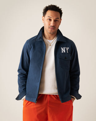 A man wearing a navy blue jacket with NY on the chest, a white shirt, and bright orange pants stands facing the camera against a light, plain background.