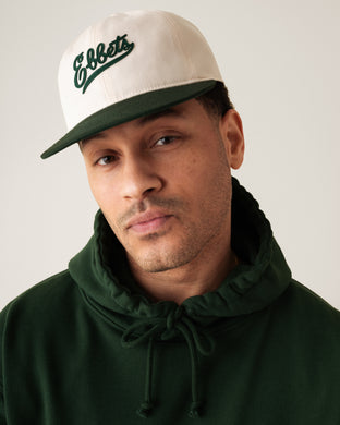 A man wearing a dark green hoodie and a white baseball cap with “Ebbets” written in green script looks at the camera against a plain background.