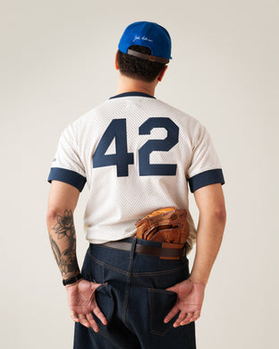 A man wearing a blue cap, white jersey with the number 42, and jeans stands facing away, hands behind his back with a baseball glove tucked into his waistband.