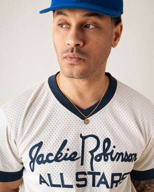 A man in a blue cap and gold necklace wears a white mesh baseball jersey featuring the text “Jackie Robinson All Stars.” He looks slightly to his left against a neutral background.