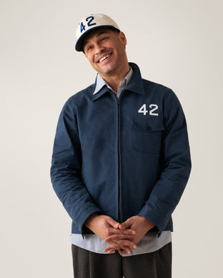 A man smiles while wearing a cream baseball cap and a navy jacket, both featuring the number 42. He stands with his hands clasped in front of him against a plain background.