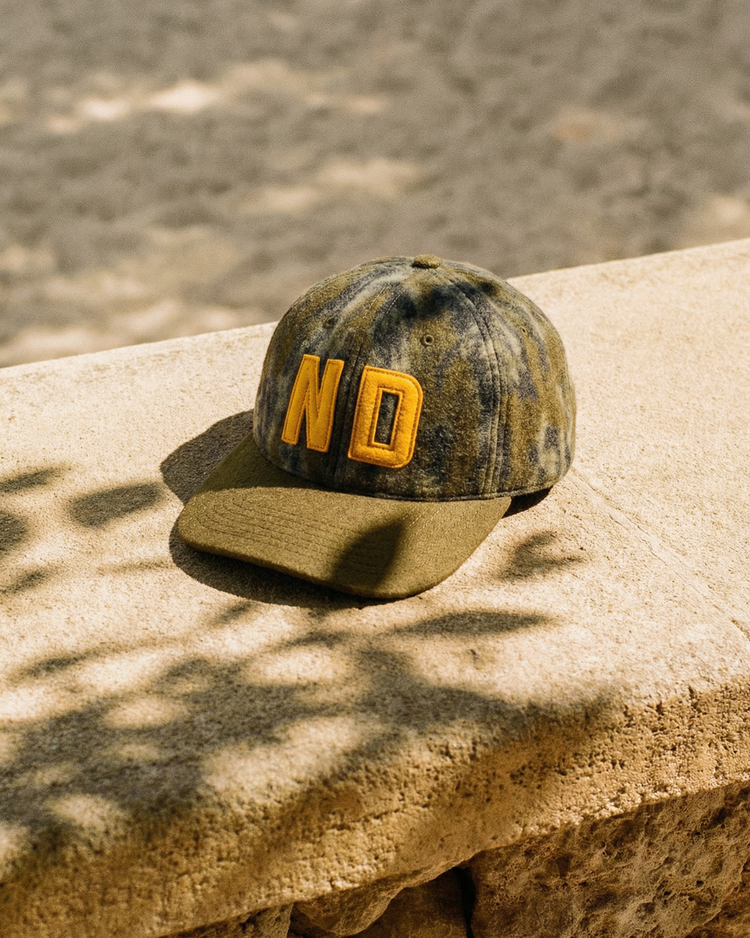 The Ebbets Field Mossy Oak x 1966 University of Notre Dame Ballcap, featuring ND letters and camo design, rests on a stone ledge in sunlight.