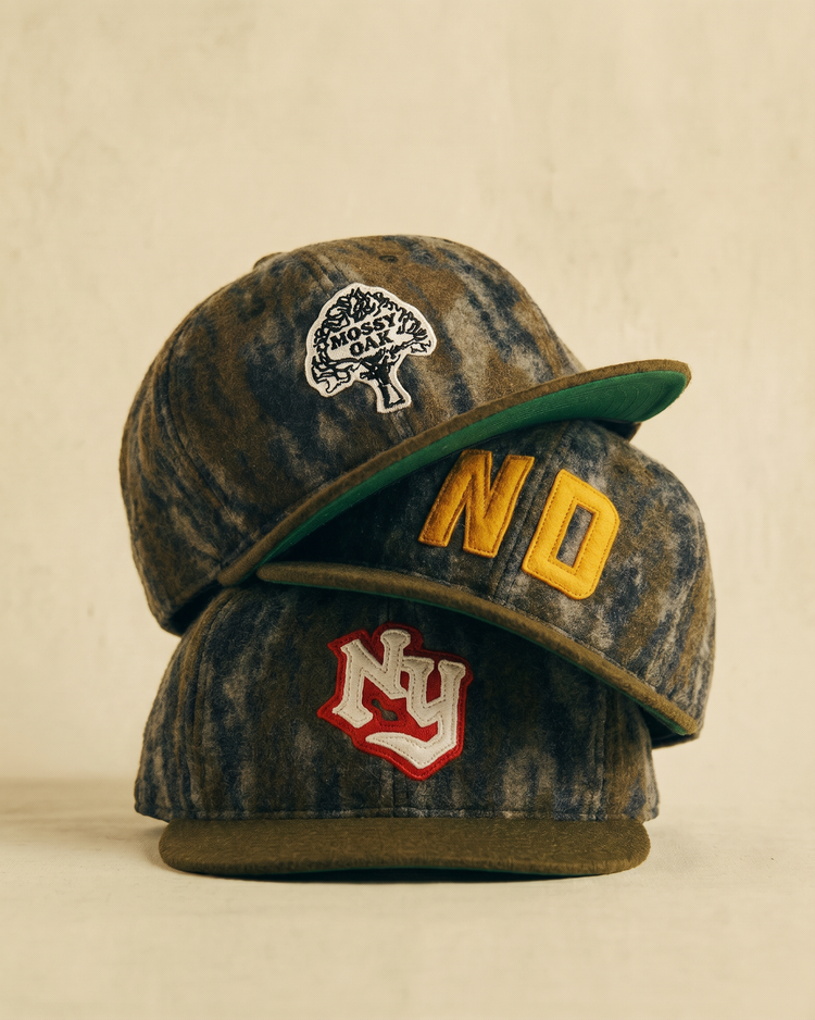 Three vintage-style Ebbets Field caps are stacked: the top is a Mossy Oak x Ebbets Field 1945 Kansas City Monarchs Ballcap, the middle has bold yellow “ND,” and the bottom shows a red and white 