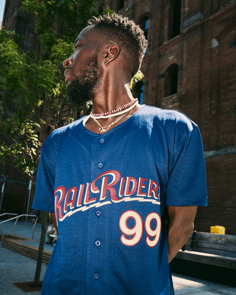 A man stands outdoors by a brick building, sunlight casting shadows as he wears an Ebbets Field Aaron Judge Scranton/Wilkes-Barre Railriders Mesh Button-Up Jersey and layered necklaces, looking to the side.