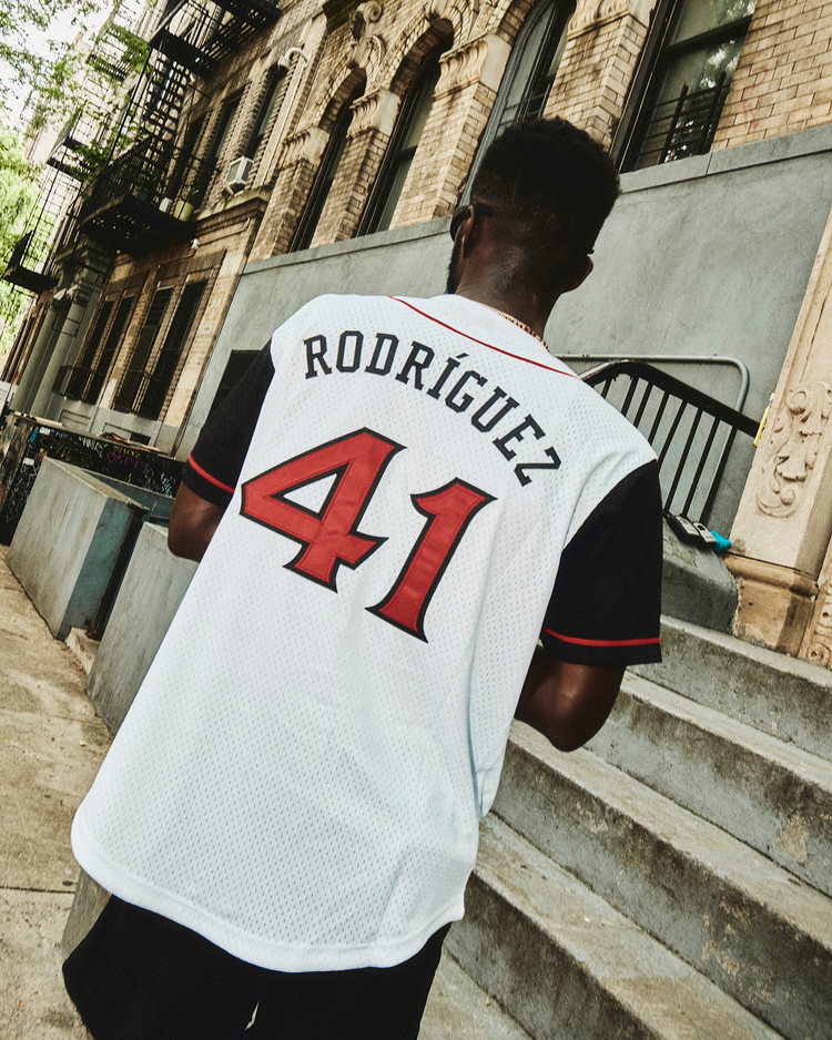 Someone wearing an Ebbets Field Julio Rodríguez Arkansas Travelers Mesh Button-Up Jersey with the red number 41 walks on a city sidewalk beside stone steps and brick buildings.