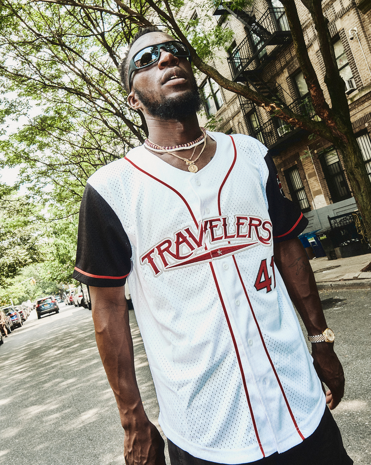 A man in sunglasses, a gold chain, and an Ebbets Field Julio Rodríguez Arkansas Travelers Mesh Button-Up Jersey stands on a tree-lined city street, looking upward with a confident expression.