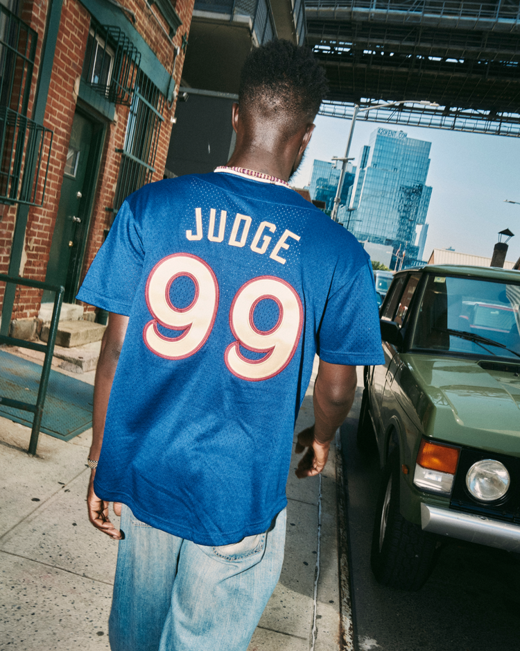 Someone wearing the Ebbets Field Aaron Judge Scranton/Wilkes-Barre Railriders Mesh Button-Up Jersey walks along a city sidewalk beside a green car, with tall buildings in the background.