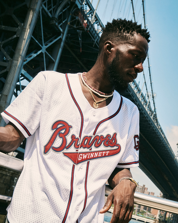 A man in an Ebbets Field Ronald Acuña Jr. Gwinnett Braves Mesh Button-Up Jersey and gold chains stands outdoors by a railing, with a large bridge and city buildings behind him on a sunny day.