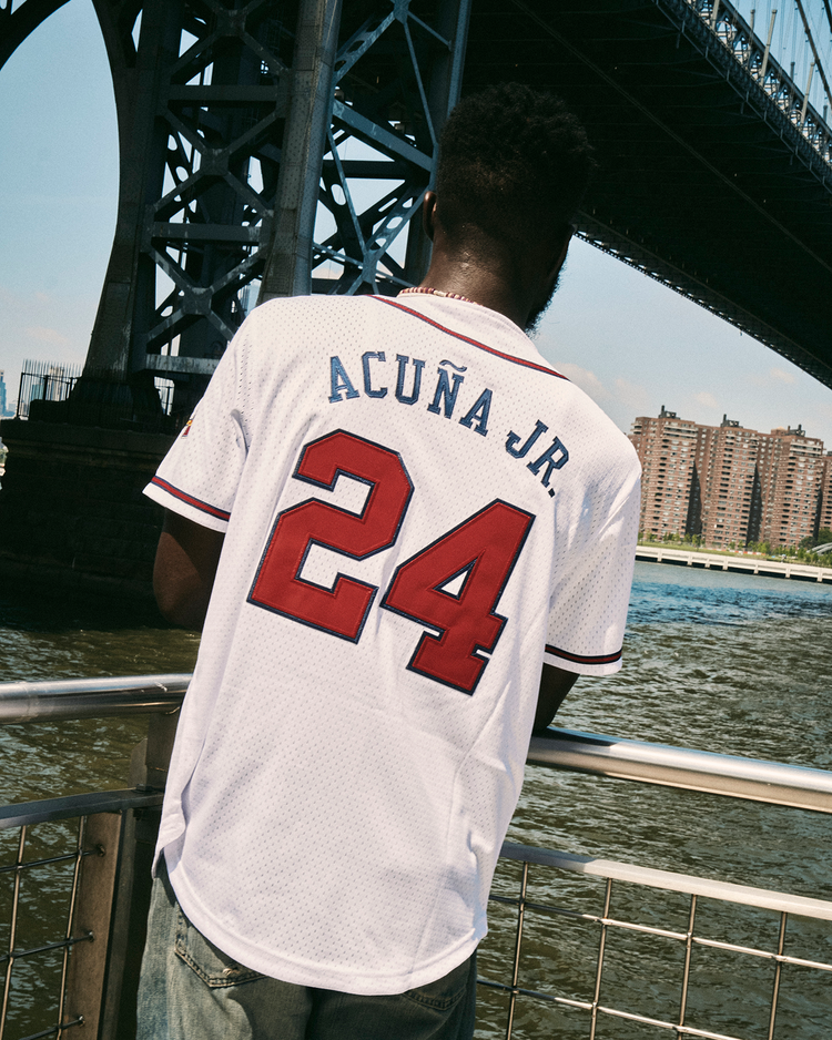 A man stands by a riverside railing under a large bridge, wearing an Ebbets Field Ronald Acuña Jr. Gwinnett Braves Mesh Button-Up Jersey. City buildings are visible across the water.