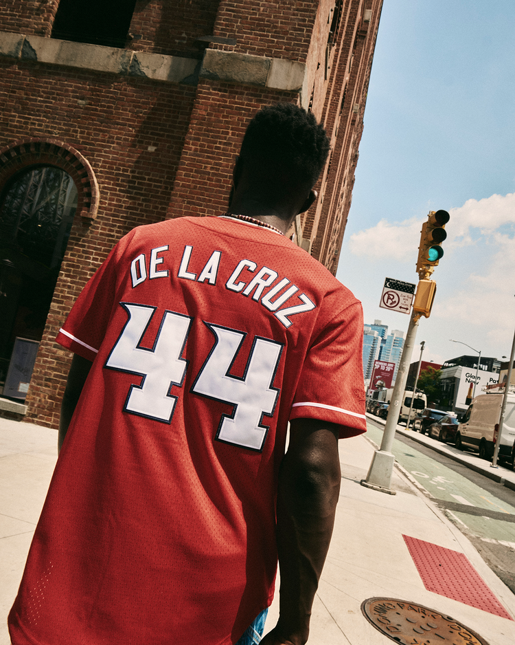 Someone in an Ebbets Field Elly De La Cruz Louisville Bats Mesh Button-Up Jersey walks along a city sidewalk by a brick building, with cars, a traffic light, and skyscrapers in the background.