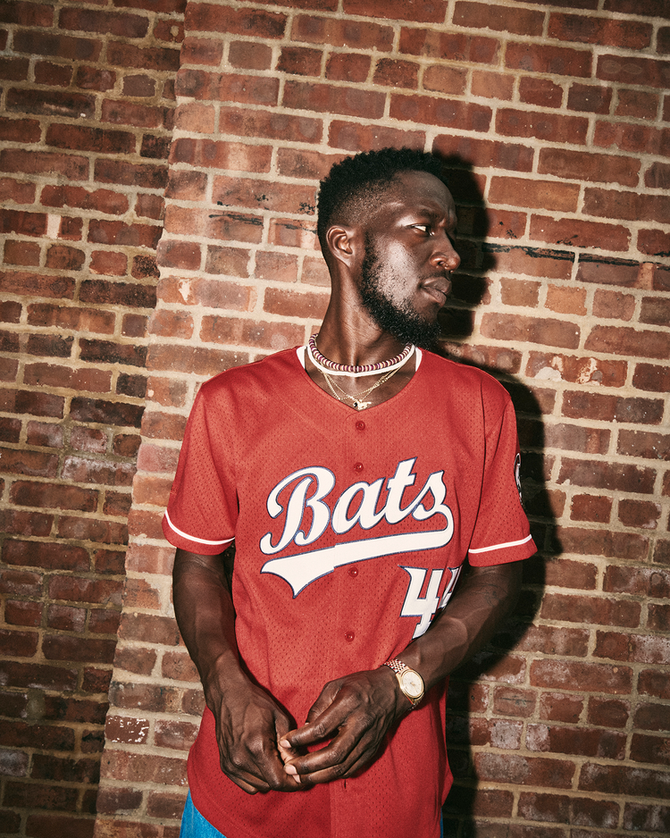 A man in an Ebbets Field Elly De La Cruz Louisville Bats Mesh Button-Up Jersey with number 44 stands by a brick wall, looking left. He sports a trimmed beard, gold chain, and wristwatch.