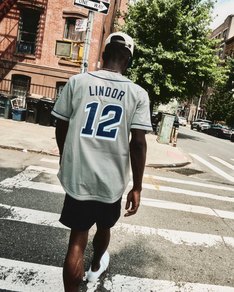 A person in an Ebbets Field Francisco Lindor Columbus Clippers Mesh Button-Up Jersey with number 12 crosses a sunny city street, with buildings and trees in the background, showcasing the throwback minor league style.