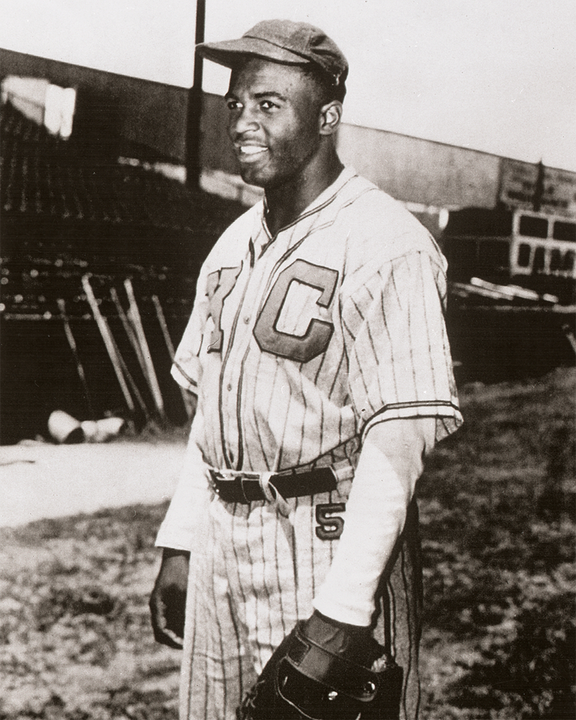 A Black baseball player in a striped uniform and cap stands on a field, smiling and holding a glove. The uniform displays large letters KC. Bleachers and baseball bats are visible in the background.