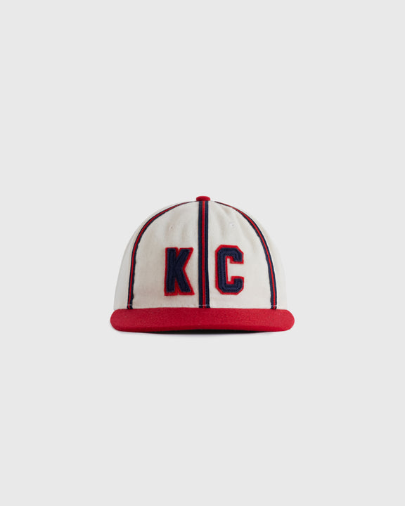 A white baseball cap with red trim and a red brim, featuring large blue-outlined red letters KC on the front.