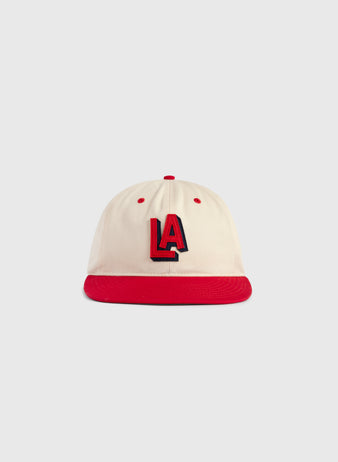 The Ebbets Field Los Angeles Angels (PCL) 1942 Logo Cotton Twill Ballcap features a cream cotton twill body, red brim and button, and bold red-and-black embroidered LA letters. Inspired by the 1942 team cap. Set on a light gray background.