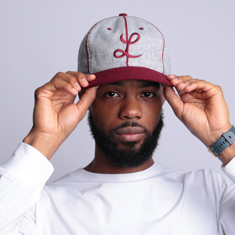 A bearded man adjusts the brim of his EFF Loyola Marymount University 1939 Vintage Ballcap—gray and maroon with a cursive L. He wears a white long-sleeve shirt, gray wristwatch, and stands against a plain light background.