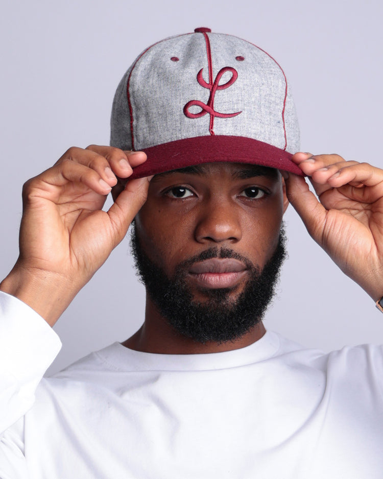 A bearded man adjusts the brim of his EFF Loyola Marymount University 1939 Vintage Ballcap—gray and maroon with a cursive L. He wears a white long-sleeve shirt, gray wristwatch, and stands against a plain light background.