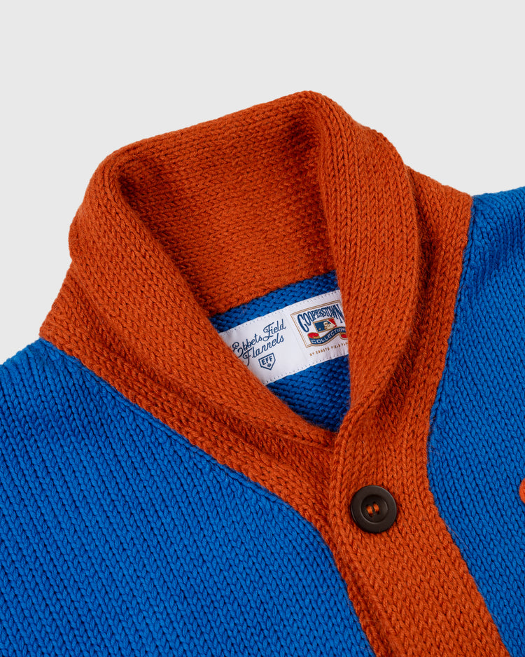 A close-up of the Ebbets Field Flannels New York Mets 1962 Wool Shawl Collar Sweater, inspired by vintage on-field styles with a burnt orange collar, bright blue body, brown buttons, and a woven label at the neckline.