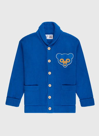 The Ebbets Field Flannels Chicago Cubs 1962 Wool Shawl Collar Sweater features wooden buttons, two front pockets, and a yellow and blue bear face patch on the left chest, inspired by a classic Cubs replica baseball sweater. Shown on a white background.