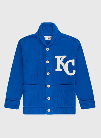 The Ebbets Field Flannels Kansas City Royals 1969 Wool Shawl Collar Sweater is a royal blue replica with tan buttons, two front pockets, a white KC patch on the left chest, and classic ribbed cuffs and hem.