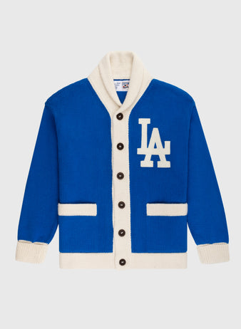 The Ebbets Field Flannels LA Dodgers 1958 Wool Shawl Collar Sweater is a blue and white varsity-style cardigan with black buttons, two front pockets, and a large LA logo on the left chest—replicating classic on-field baseball sweaters.