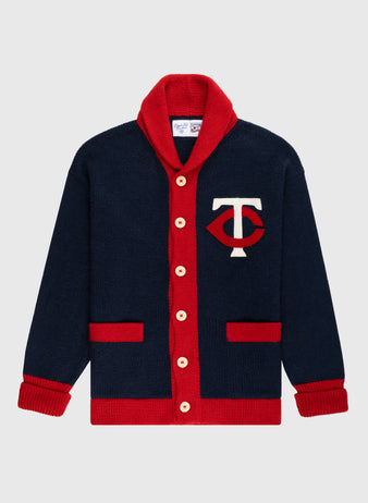 The Ebbets Field Flannels Minnesota Twins 1961-71 Wool Shawl Collar Sweater features navy wool, red trim, cream buttons, two red pockets, and a white T with a red eye-shaped design—perfect for true Twins fans seeking vintage style.