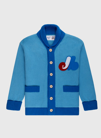 The Montreal Expos 1969 Wool Shawl Collar Sweater by Ebbets Field Flannels is a blue cardigan with darker blue trim, beige buttons, two front pockets, and a red, white, and blue chest patch inspired by classic baseball sweaters.