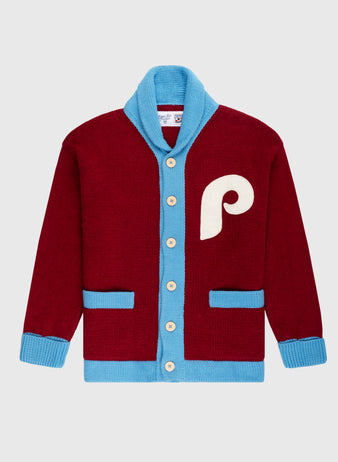 The Philadelphia Phillies 1979 Wool Shawl Collar Sweater by Ebbets Field Flannels features maroon wool, blue trim, cream buttons, blue cuffs and pockets, plus a large cream P on the chest—replicating classic on-field baseball sweaters. Light gray background.