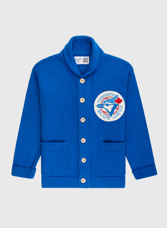 The Ebbets Field Flannels Toronto Blue Jays 1977 Wool Shawl Collar Sweater features royal blue knit, white buttons, two front pockets, and a large team logo patch on the left chest—styled after vintage on-field baseball sweaters.