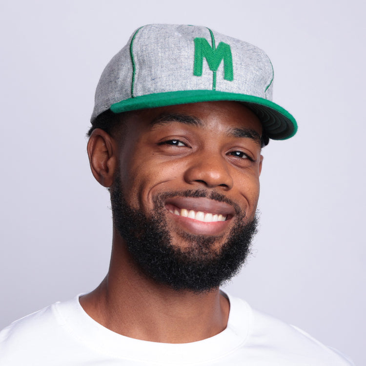 A smiling man with a short beard wears the EFF Manhattan College 1932 Vintage Ballcap—gray wool baseball cloth with a green M—paired with a white shirt, standing against a light gray background.
