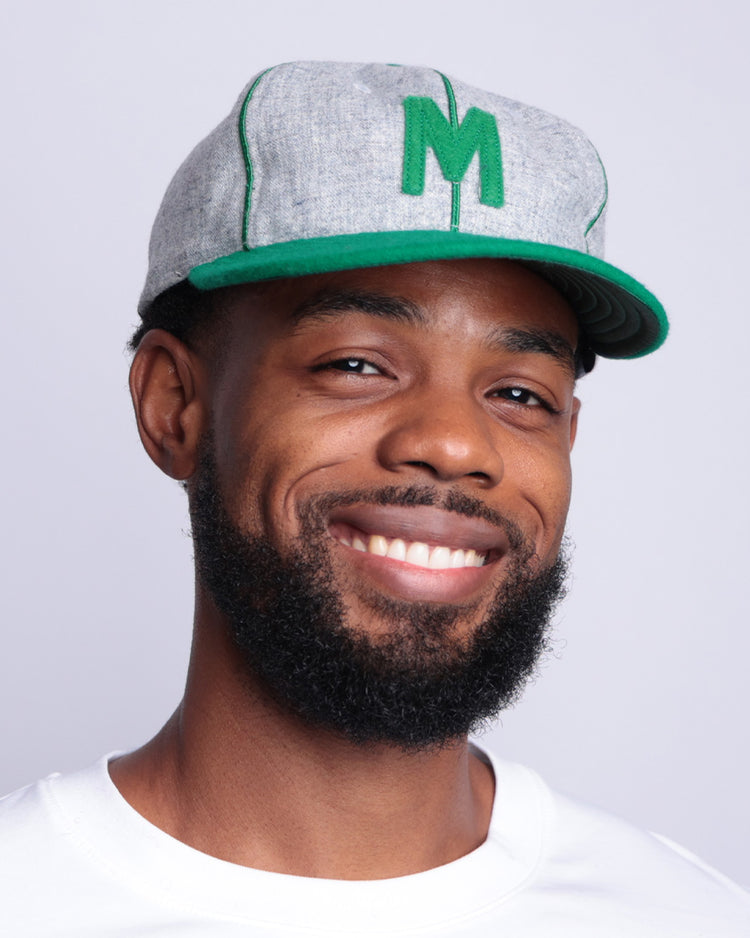 A smiling man with a short beard wears the EFF Manhattan College 1932 Vintage Ballcap—gray wool baseball cloth with a green M—paired with a white shirt, standing against a light gray background.